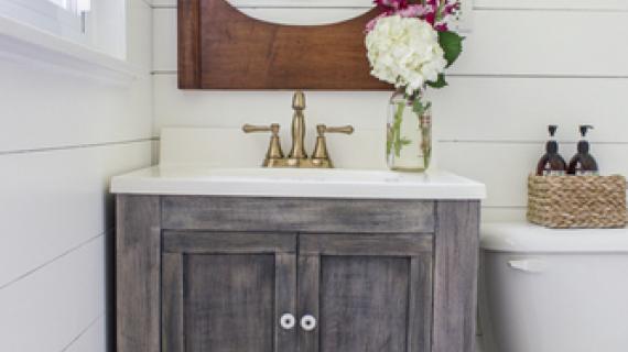 gray stained small bath vanity with turned legs and an open shelf with basket in white bathroom